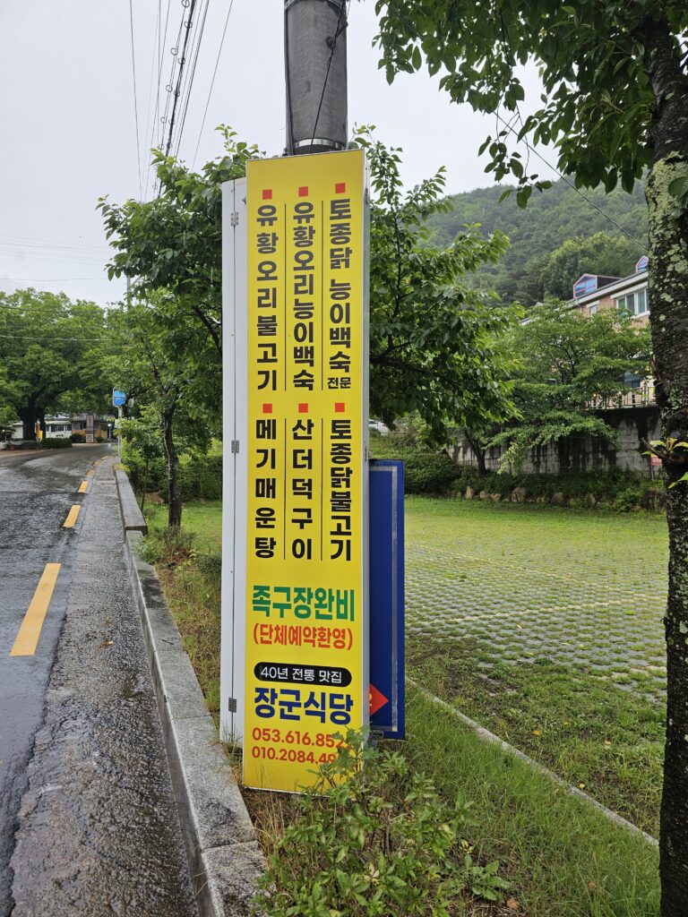 Yellow Janggun Sikdang roadside sign beside a wet road in Dalseong-gun, Daegu.