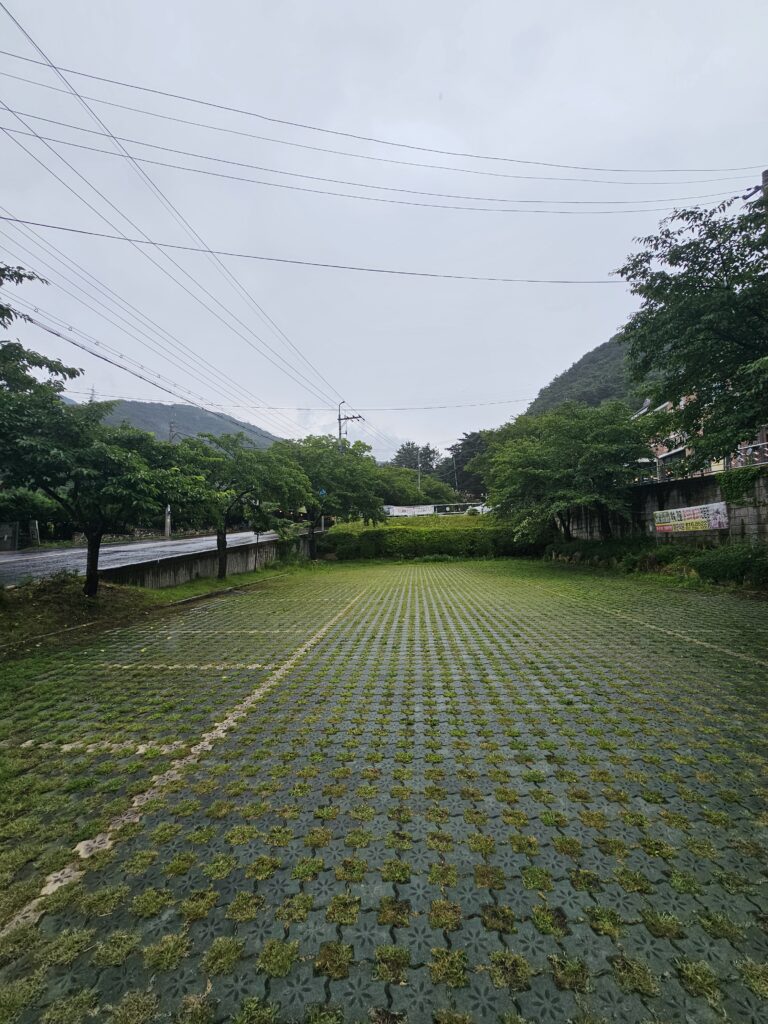 Wide view across Janggun Sikdang parking lot toward trees, road, and hills.