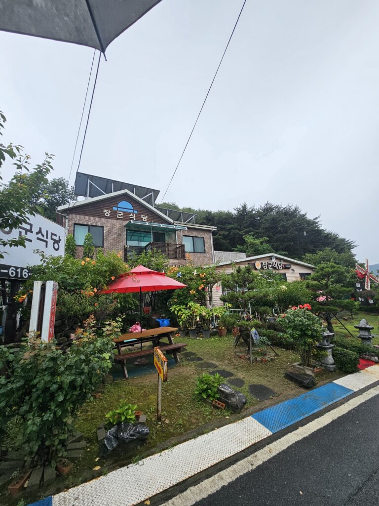 Janggun Sikdang exterior with red umbrella, garden tables, potted plants, and flowers.
