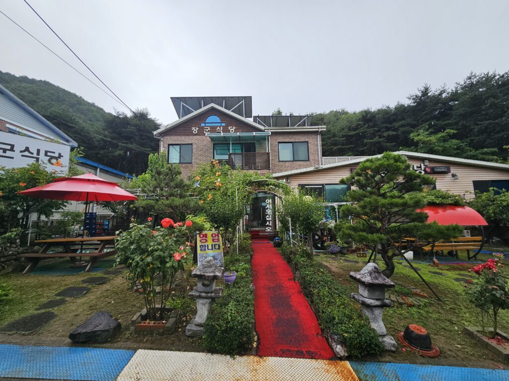 Red entrance path between flowers and stone lanterns at Janggun Sikdang on a rainy day.