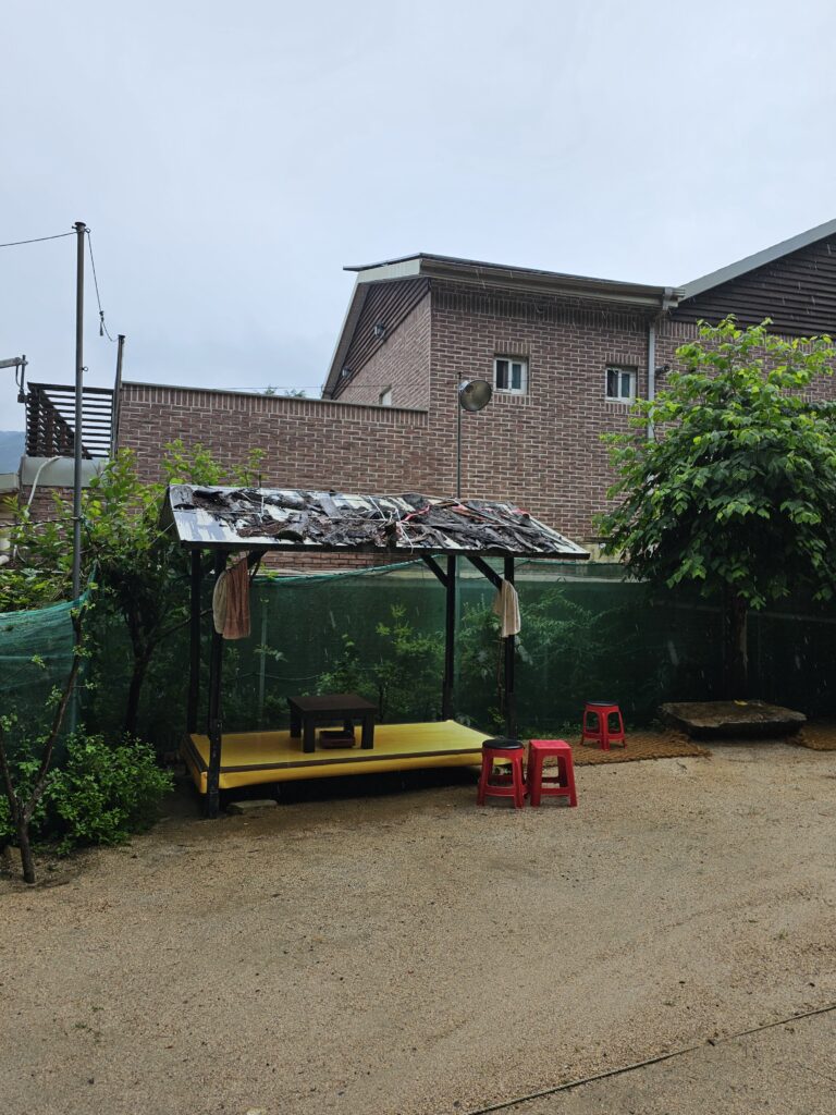 Small covered yellow platform with low table and red stools beside the jokgu court.