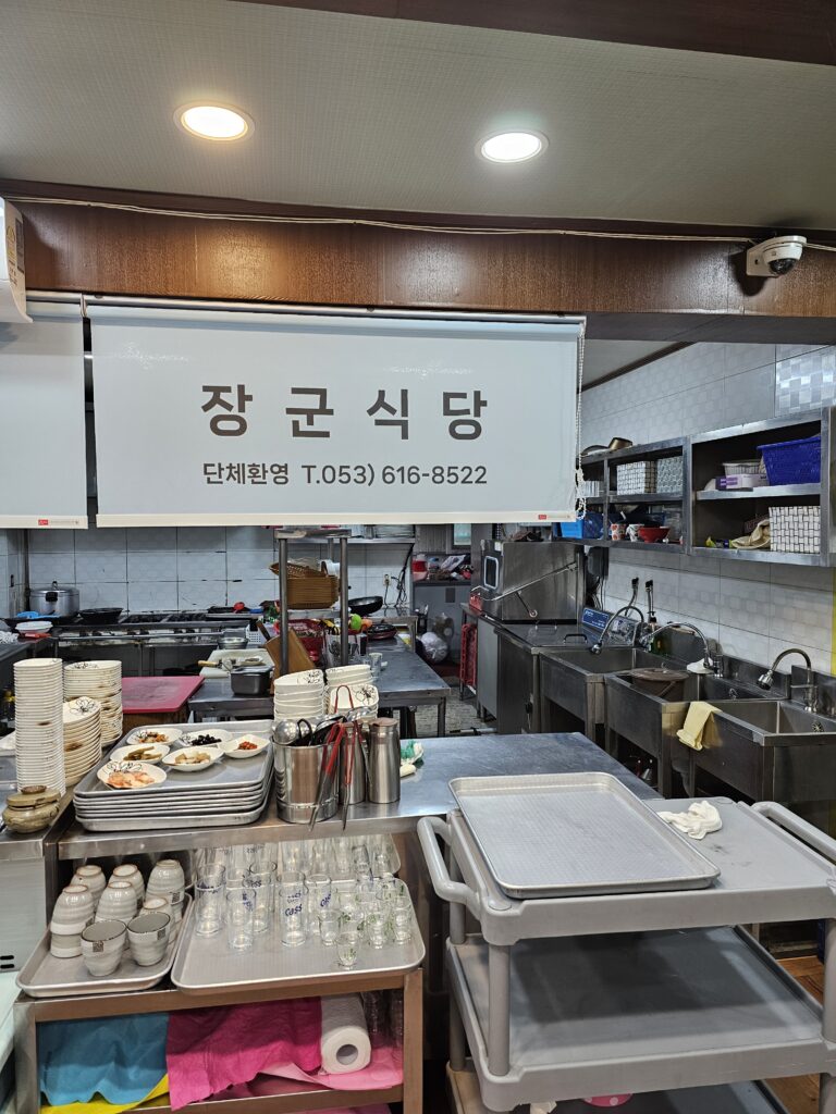 Open kitchen at Janggun Sikdang with sinks, worktables, dishes, trays, and restaurant sign.