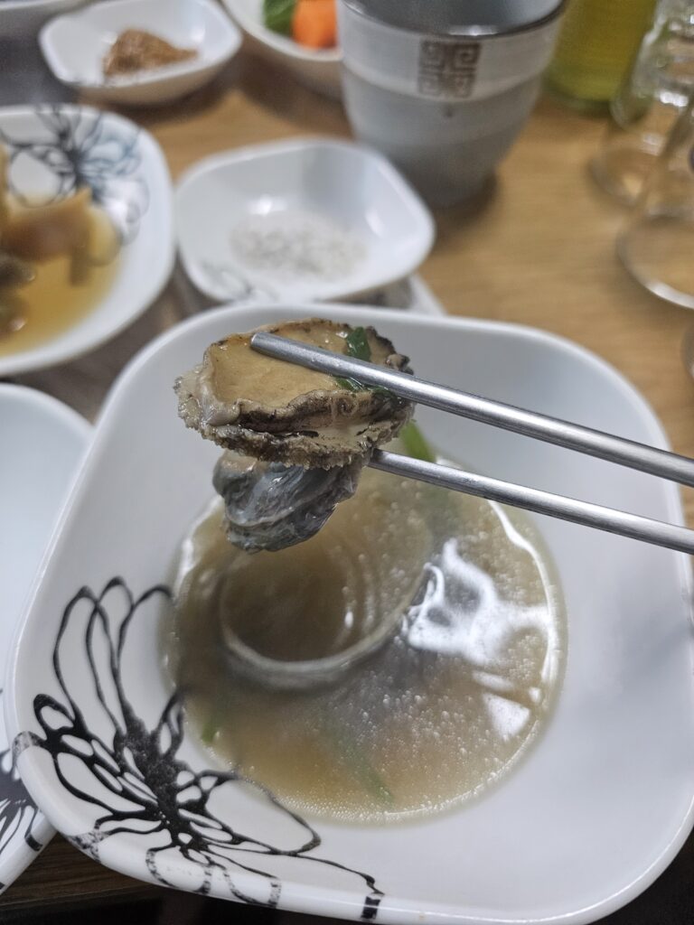 Abalone in its shell held with chopsticks above a bowl of clear broth.