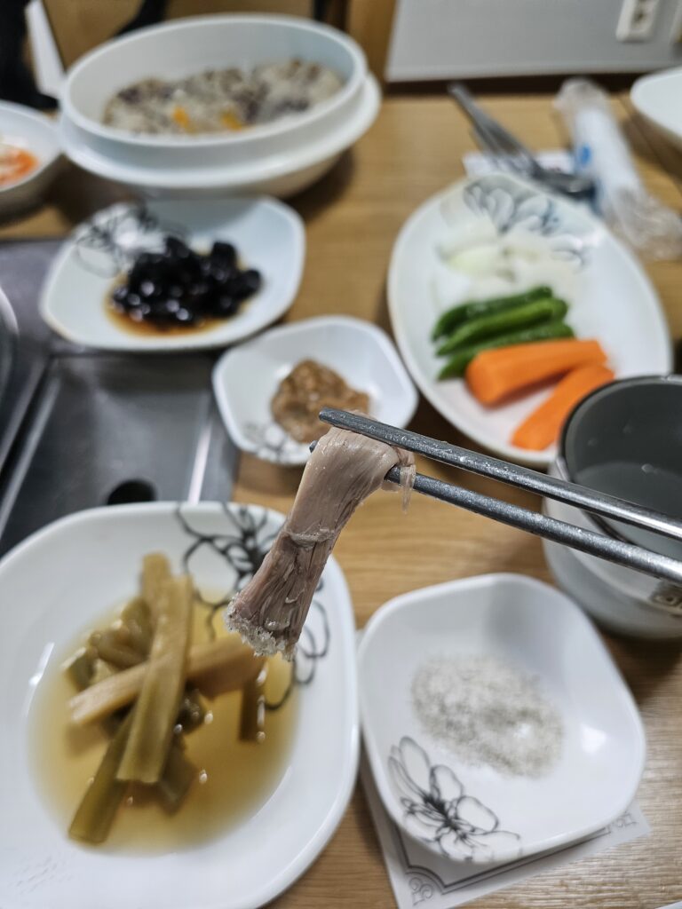 Close-up of a piece of duck meat held with chopsticks above Korean side dishes.