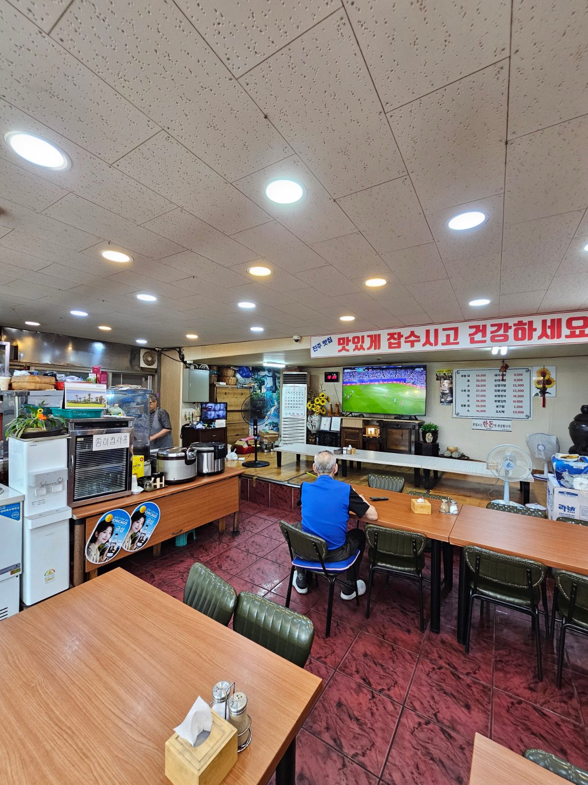 Simple dining room with tables, wall menu, and TV at Jinju Wang Bossam
