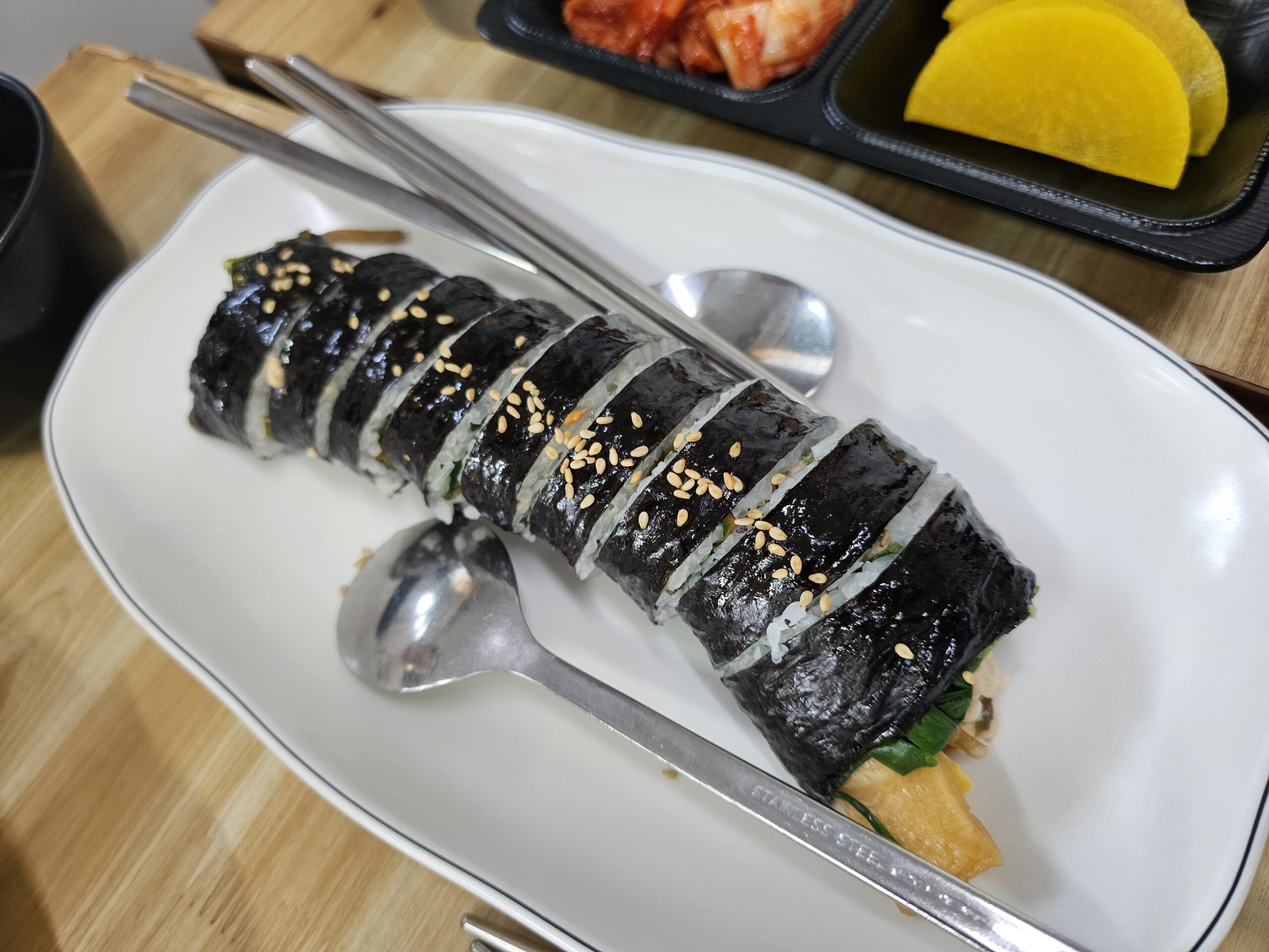 ゴマをのせて切り分けたツナキンパ、スプーン、箸、キムチ、たくあんと一緒に