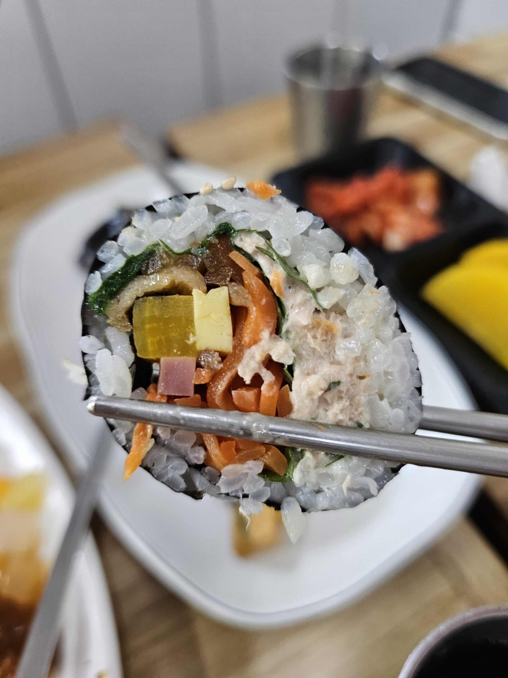 箸でつまんだツナキンパ1切れ。たくあん、にんじん、野菜が見える