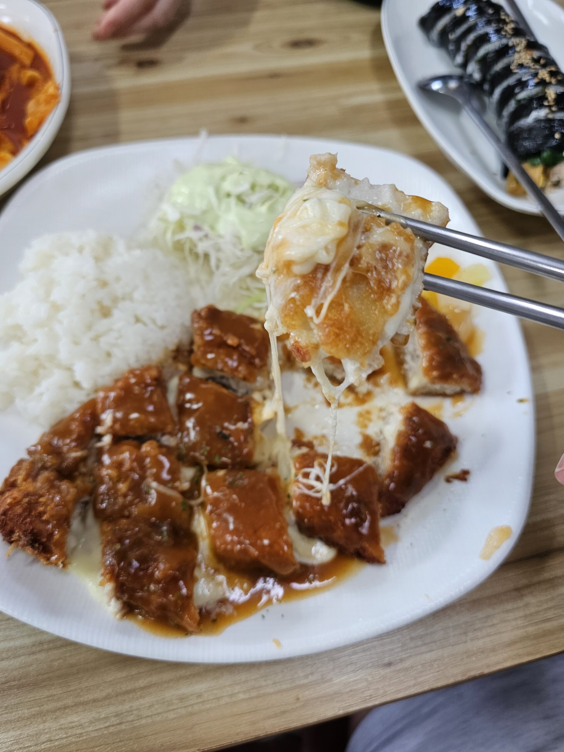 ご飯、キャベツ、たくあんの上で箸で持ち上げたチーズトンカツ一切れ